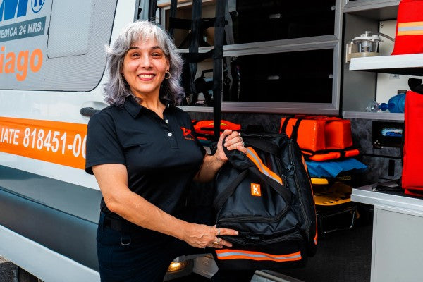 Fotografía de una mujer afuera de una ambulancia sosteniendo un botiquín de equipo para paramédicos color negro con naranja.