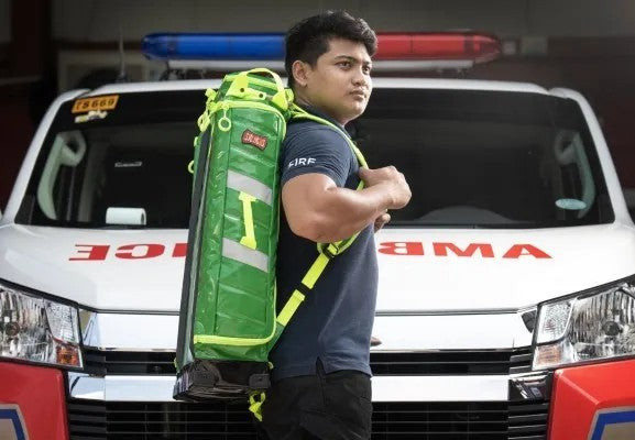 Fotografía de un hombre que es parte de una brigada de primeros auxilios de pie portando una mochila color verde.