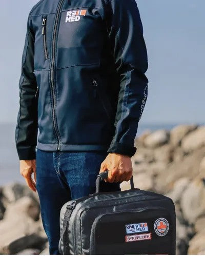 Fotografía de un hombre de pie cargando en una mano una mochila color negro que contiene un kit de emergencia.