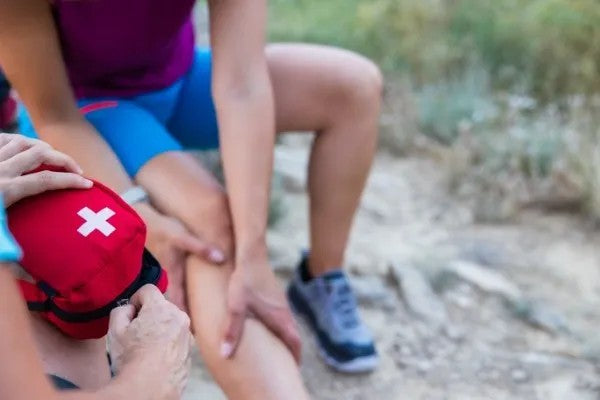 Fotografía de una persona sosteniendo su pierna mientras recibe primeros auxilios básicos y otra persona abriendo un maletín.