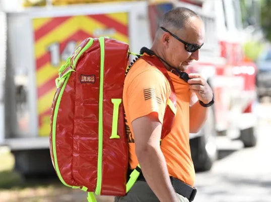 Fotografía de un hombre de camisa color naranja portando una mochila de primeros auxilios en color rojo.