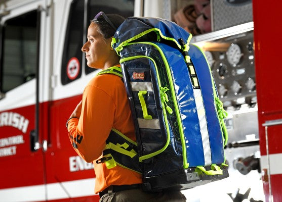 Fotografía de una mujer cargando una mochila de primeros auxilios color azul con cierres color verde.