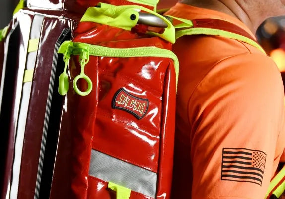 Fotografía de cerca de lado de una persona de camisa naranja portando una mochila de primeros auxilios color rojo.